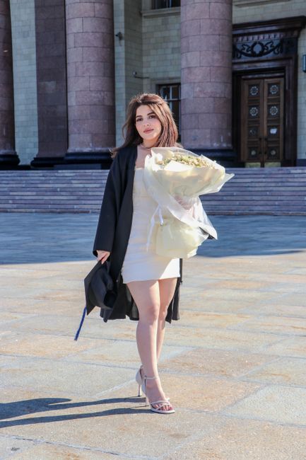 A young woman standing outdoors in front of a stone building with steps, holding a large bouquet of cream and pale yellow flowers wrapped in light-colored paper. She is dressed in a white dress with a