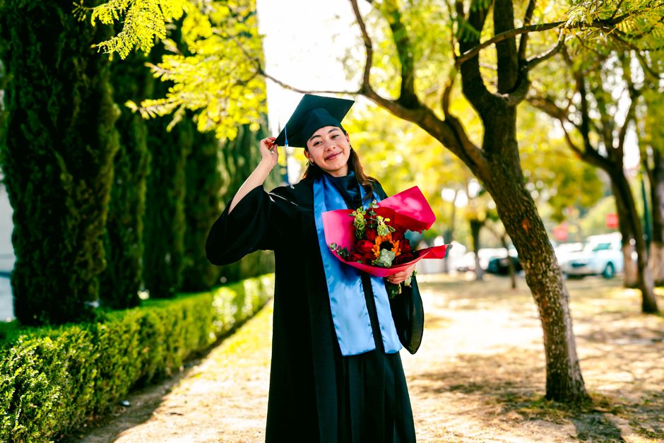 A young female graduate in a black cap and gown, wearing a blue sash, stands outdoors on a sunny day amongst green trees and neatly trimmed bushes. She is smiling and holding a vibrant bouquet wrapped