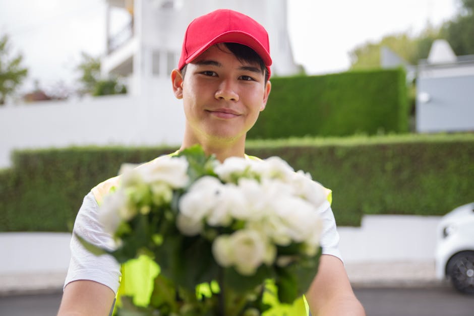 A delivery person wearing a red cap and a high-visibility vest is standing outdoors with a slight smile, holding a bouquet of white flowers closely towards the camera. The bouquet features lush, fresh