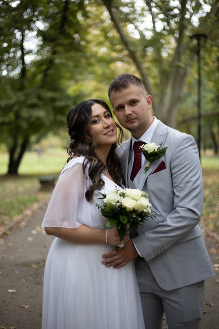 A newlywed couple stands together in a park setting during daytime, with tall trees and a pathway visible in the background. The bride, on the left, has long, wavy dark hair and is wearing a flowing w