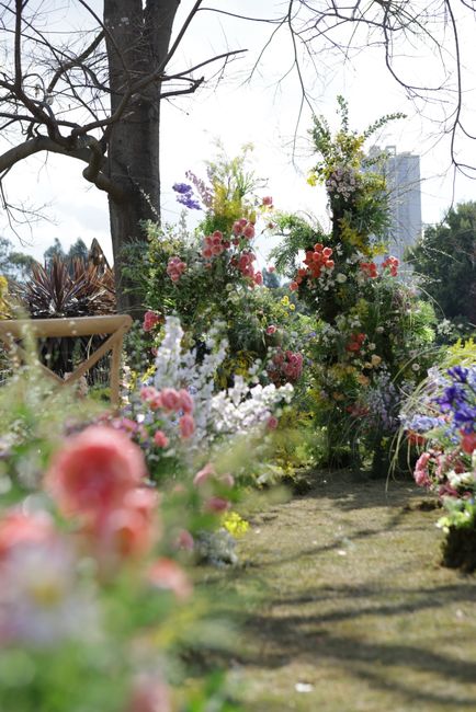 An outdoor floral display featuring a variety of fresh flowers arranged in bouquets and standing on the ground, set against a backdrop of trees with bare branches and a bright, sunlit sky. The arrange